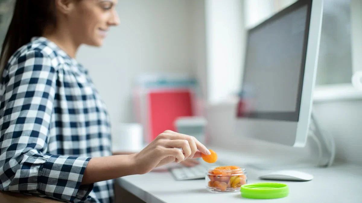 A woman smiling at her desk while picking a dried apricot from a container of office pantry snacks.